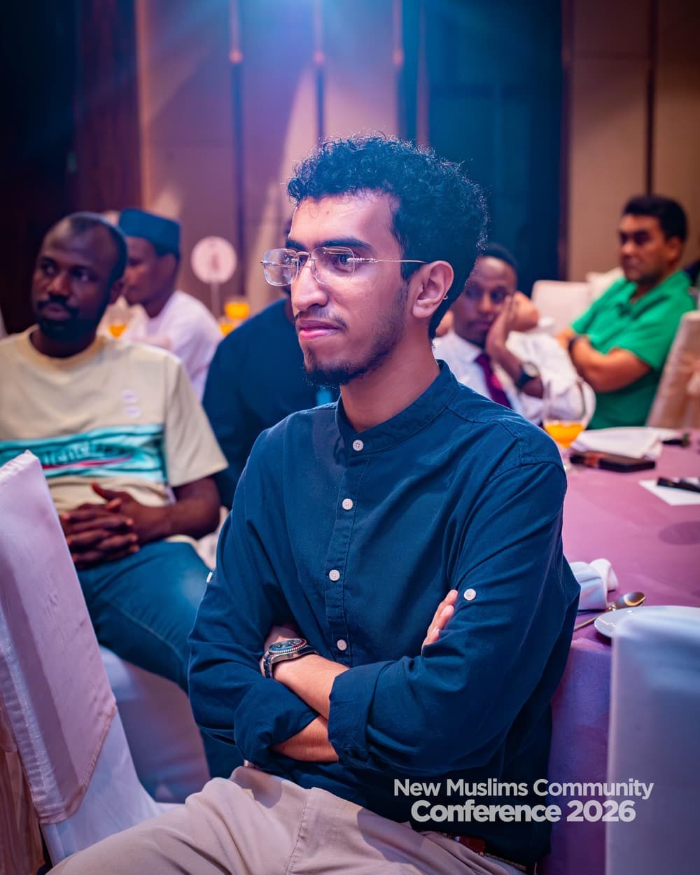 A young man with glasses listening intently from the audience, arms folded, others seated behind him