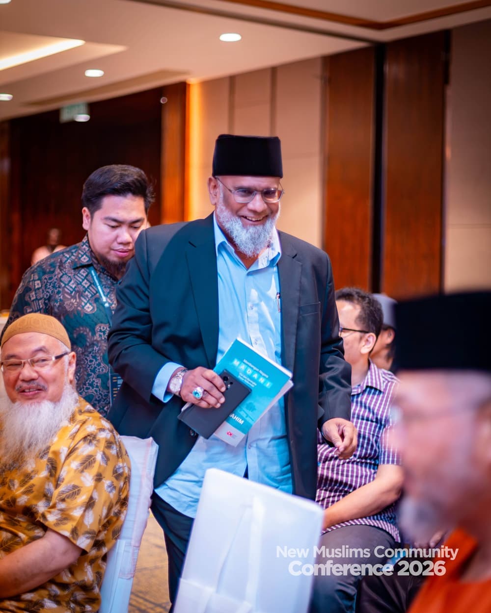 A bearded man in a songkok smiling as he walks through the hall holding a book, attendees seated around him