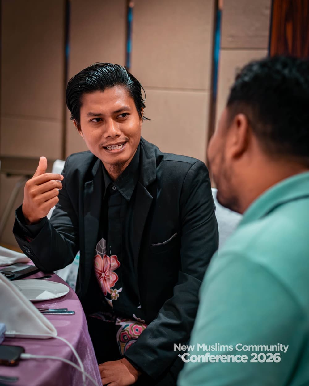 A young man in a black blazer smiling and gesturing mid-sentence to the attendee across the table