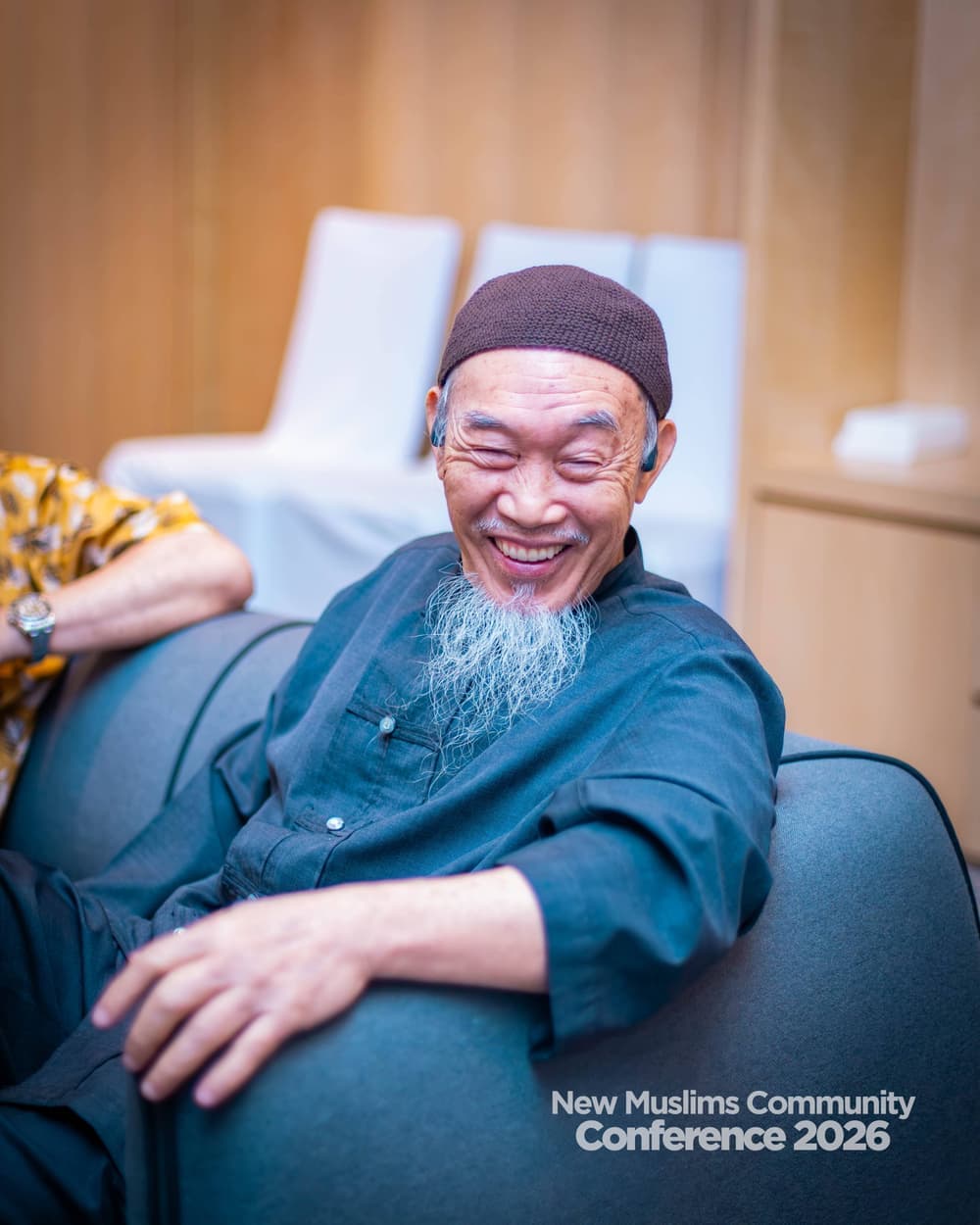 An elderly man in a black kufi with a white beard, laughing warmly while seated in an armchair