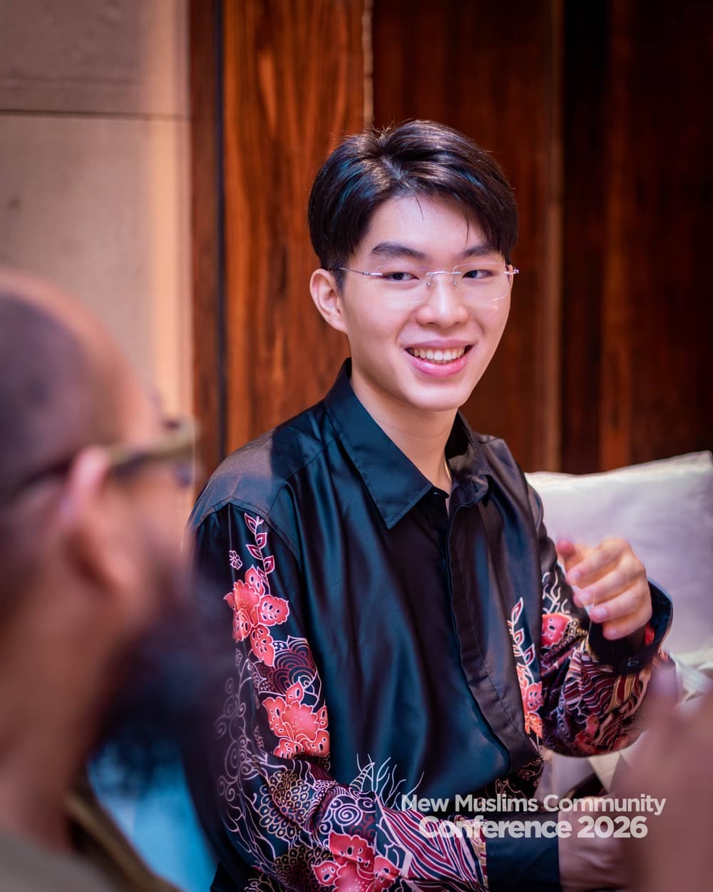 A young man in floral batik smiling as he listens to the person seated opposite him