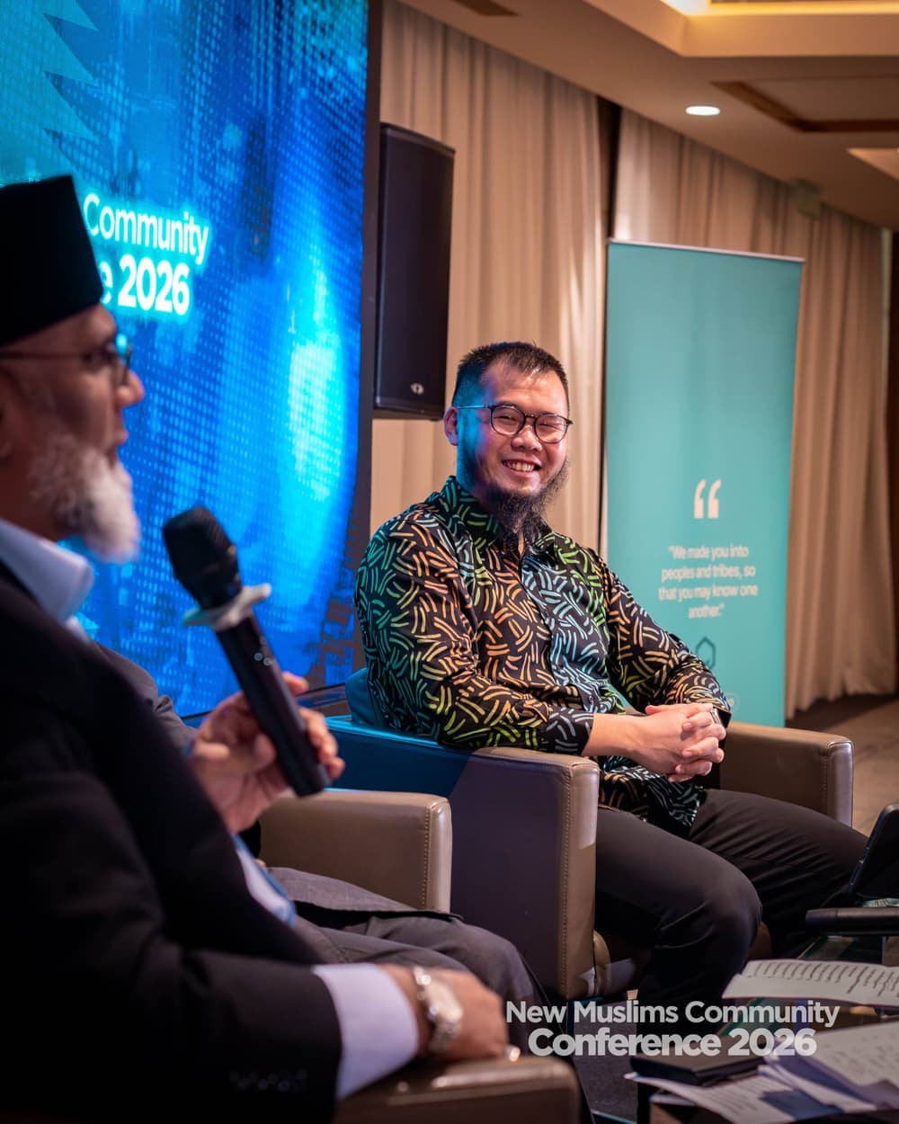 A panel speaker smiling on stage as an elder in the foreground raises a microphone to ask a question