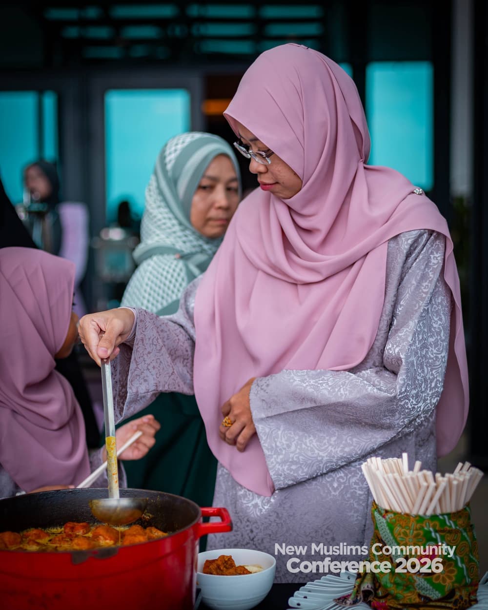 A woman in a pink hijab serving from a pot at the buffet table, two others working beside her