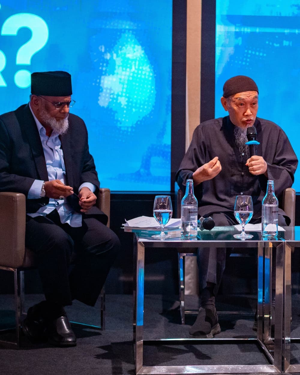 Two panellists seated on stage, one speaking into a microphone, beneath a screen reading Who Profits from Our Fear?
