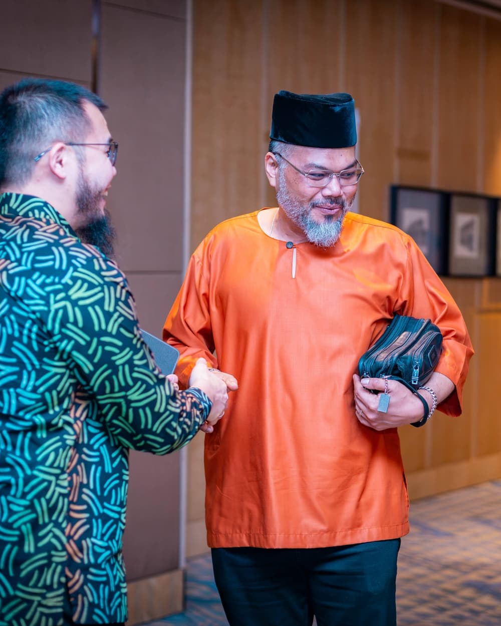 Two men shaking hands in greeting in the hallway, one in green batik and one in an orange baju melayu